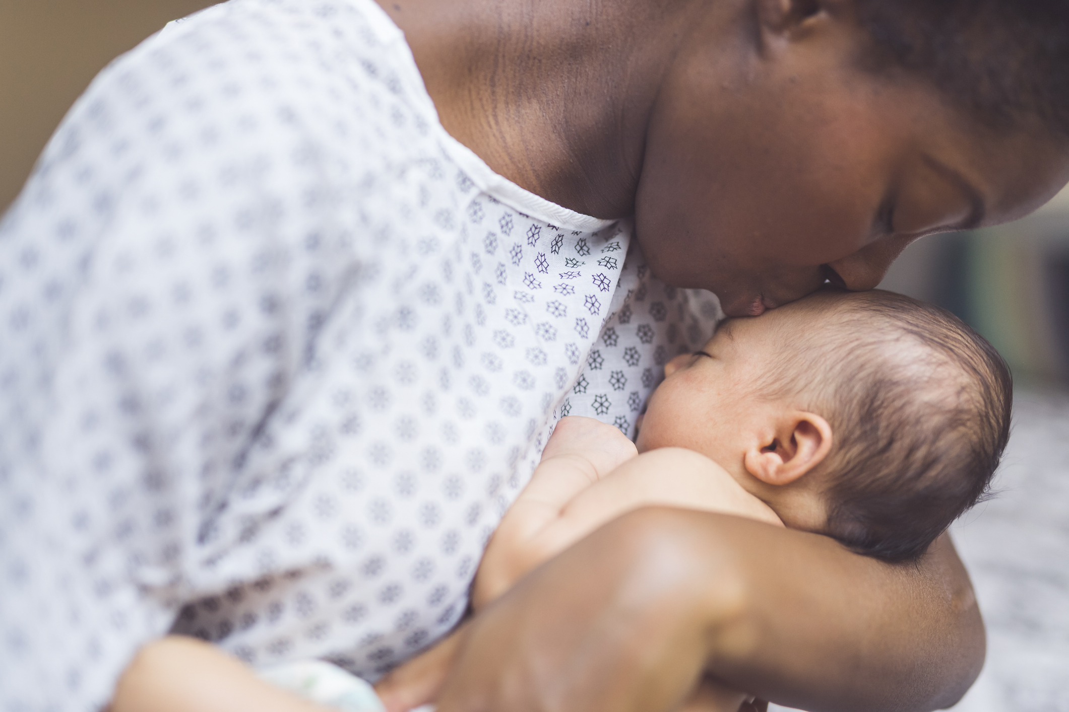 Eine junge afroamerikanische Mutter im Krankenhauskittel hält ihr Kleinkind sanft in den Armen und lächelt auf es herab. Die Augen des Babys sind geschlossen.