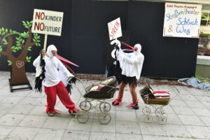 zwei Schauspieler im Storchkostum mit zwei Plakaten in der Hand. Plakat 1: No Kinder No Future; Plakat 2: Streik