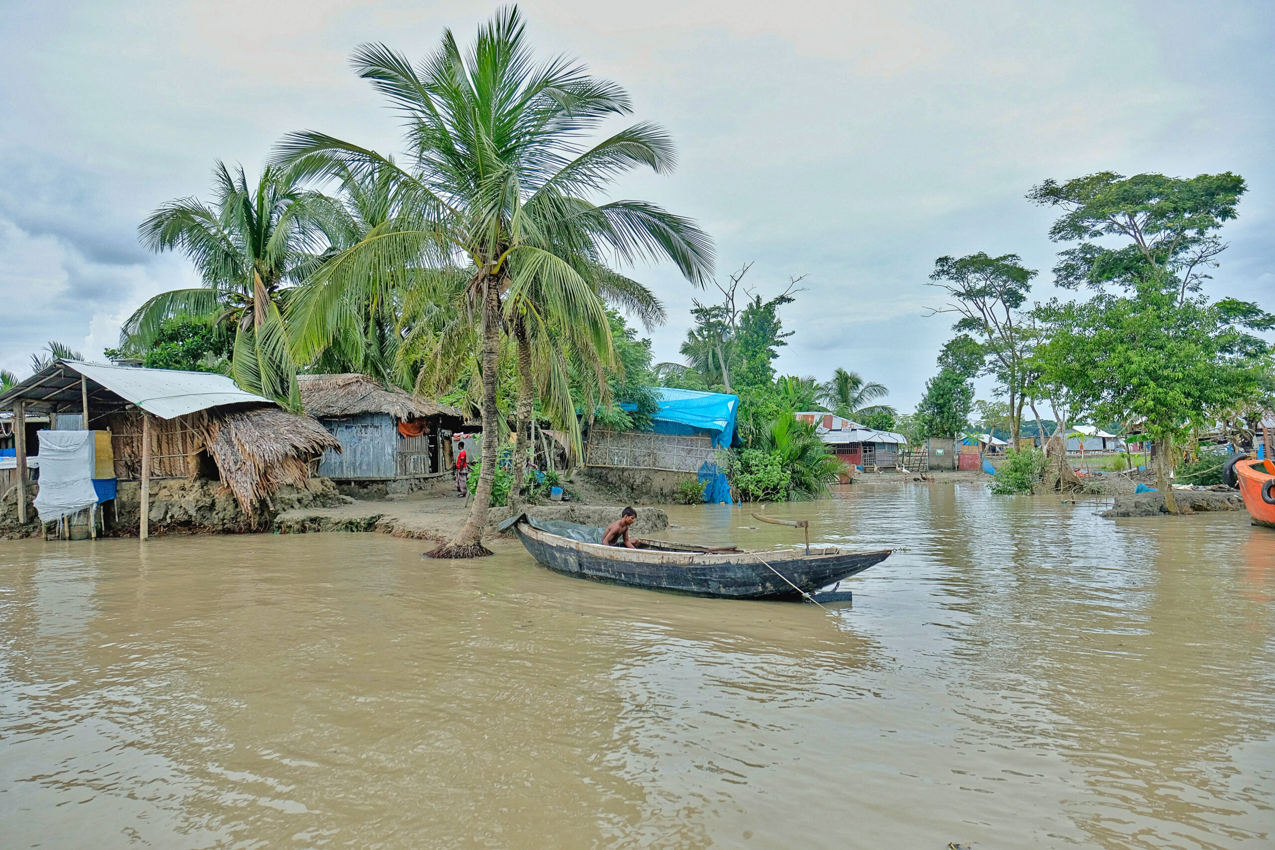 Boot auf dreckigem Gewässer in Bangladesch, Hütte und Palmen im Hintergrund.