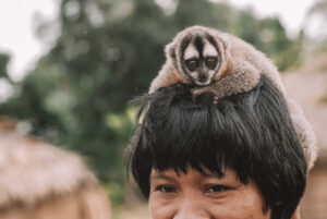 Ein kleiner Affe sitzt auf dem Kopf einer indigenen Frau im brasilianischen Amazonasgebiet. Im Hintergrund sind unscharf Bäume und Hütten zu sehen.