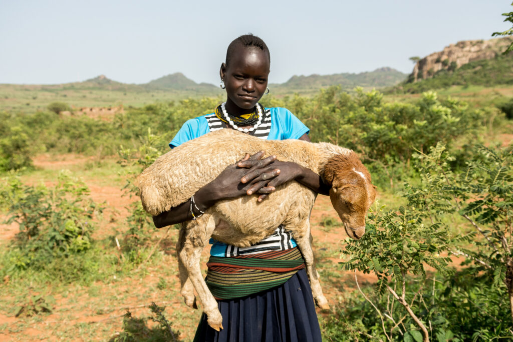 Eine Person der Karamojong steht in einer ländlichen Landschaft im Osten Ugandas und hält ein Schaf auf dem Arm. Im Hintergrund sind Büsche, rote Erde und Hügel zu sehen.