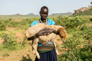 Eine Person der Karamojong steht in einer ländlichen Landschaft im Osten Ugandas und hält ein Schaf auf dem Arm. Im Hintergrund sind Büsche, rote Erde und Hügel zu sehen.