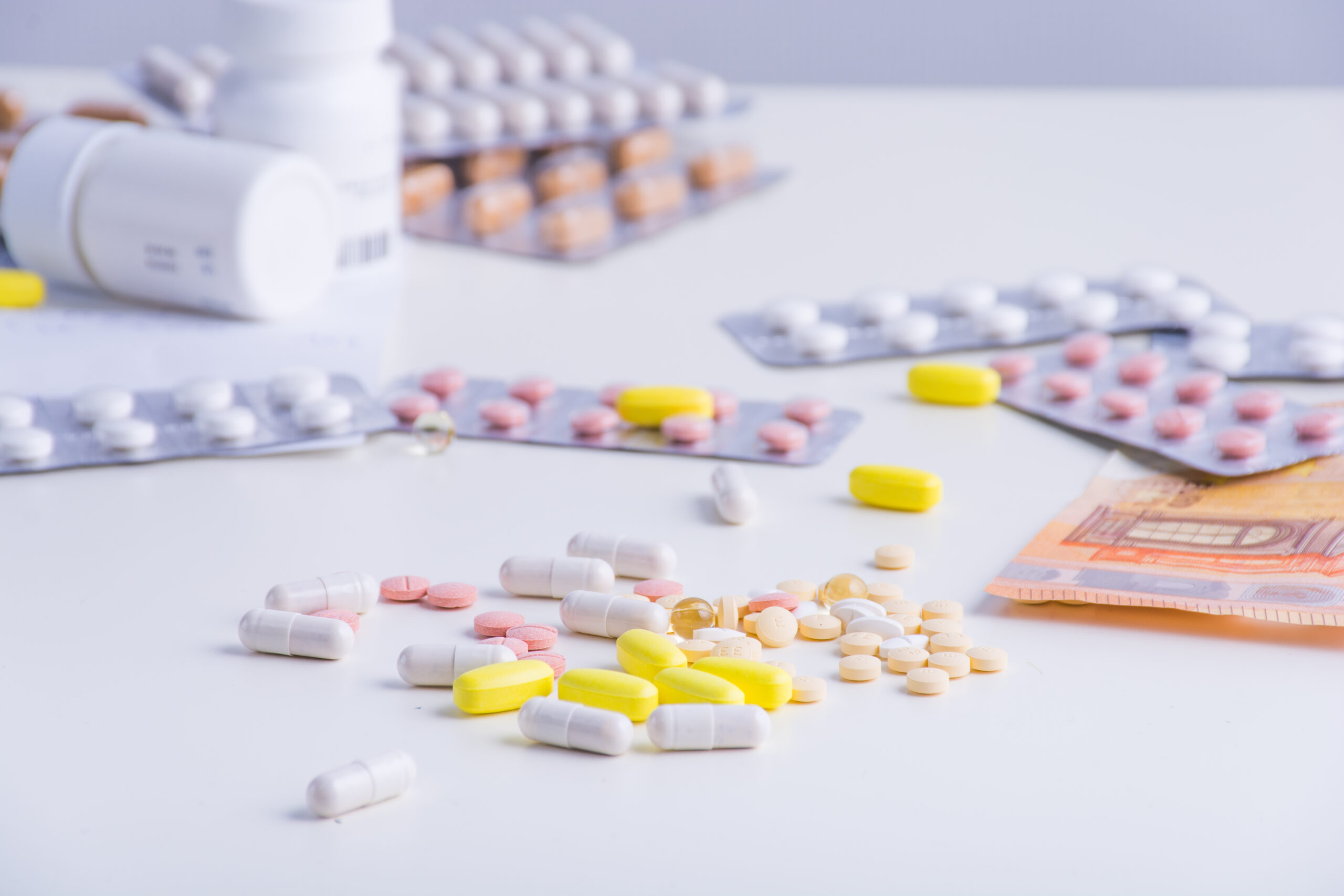 Verschiedene Tabletten und Kapseln liegen neben Medikamentenpackungen und Euro-Geldscheinen auf einer weißen Fläche.
